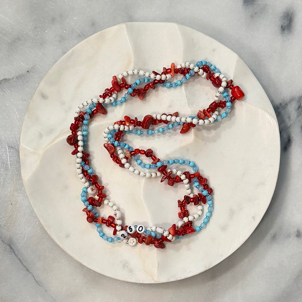 Americana Collection, 250 Anniversary, Three strand braided beaded necklace with red, white, and blue beads on a marble surface, gold stars and numbers 250, Amelia Heir Art