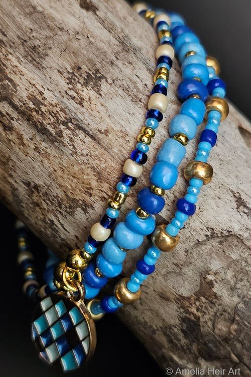 Stack of blue and cream beaded bracelets with gold accents and an ornament charm on a wooden surface