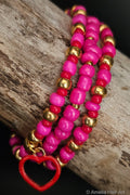 Stack of pink, red, and gold beaded bracelets with a red heart charm on a wooden surface.