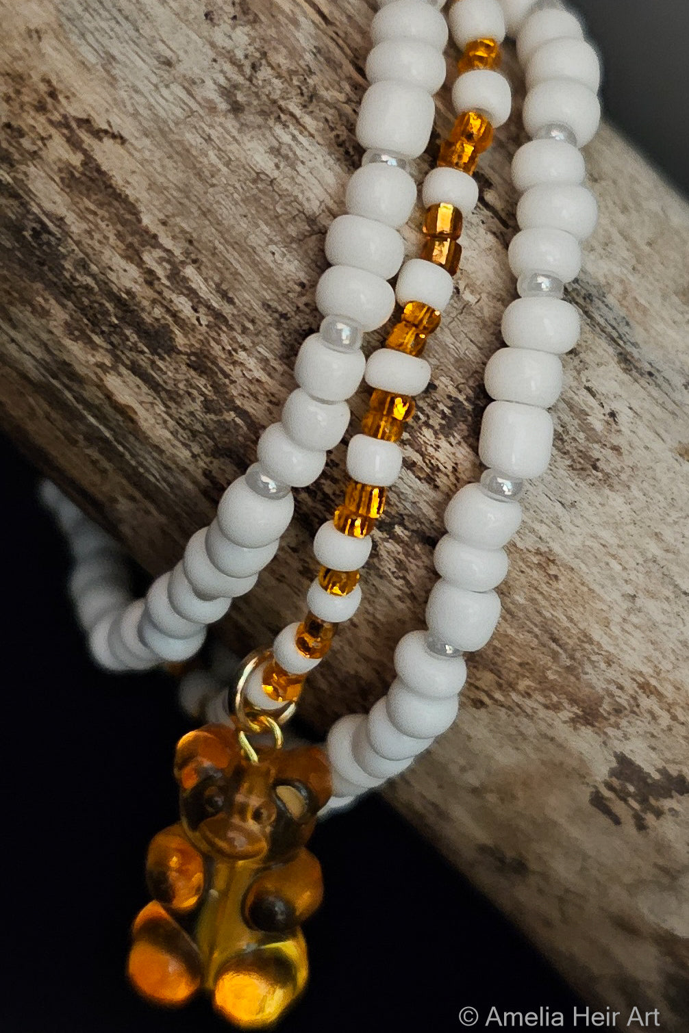 Three beaded bracelets with white beads and orange accents with a gummy-bear charm on a wooden surface.
