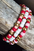Three beaded bracelets with red, pink, white, and gold beads on a wooden surface.