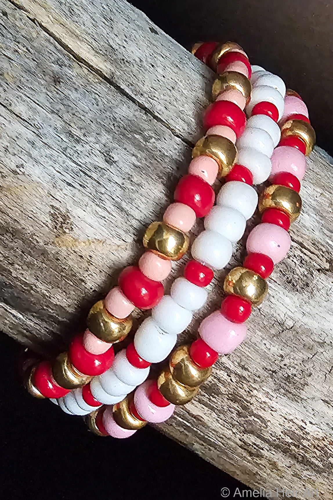 Three beaded bracelets with red, pink, white, and gold beads on a wooden surface.