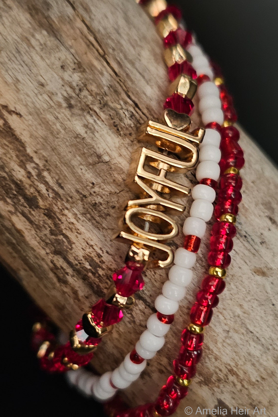 Three red and white beaded bracelets with gold accents and 'JOYFUL' charm on a wooden surface.