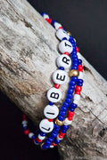 Three beaded bracelets with 'LIBERTY' on them, one red, white, and blue, the other with gold beads, on a wooden surface.