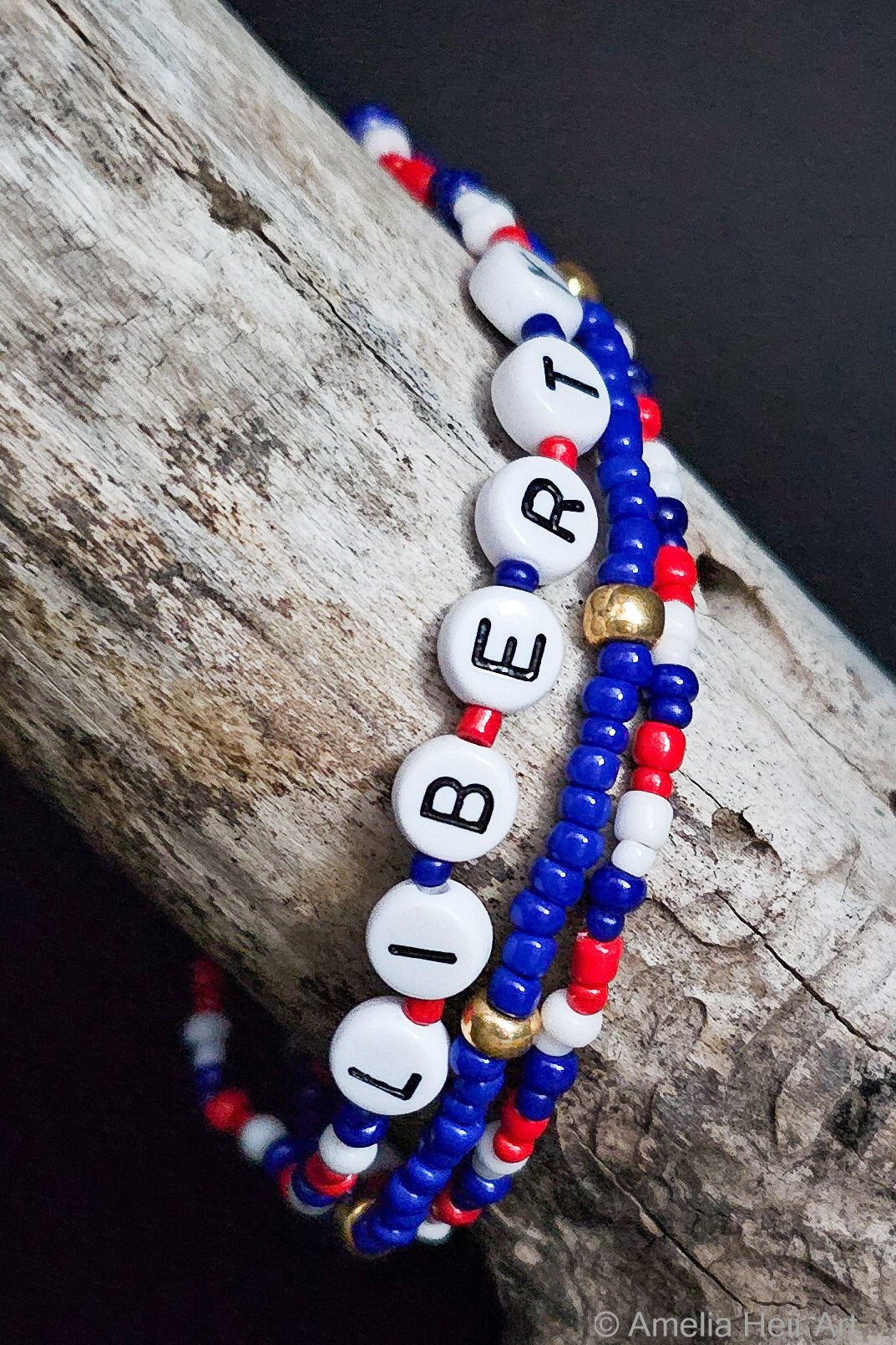 Three beaded bracelets with 'LIBERTY' on them, one red, white, and blue, the other with gold beads, on a wooden surface.