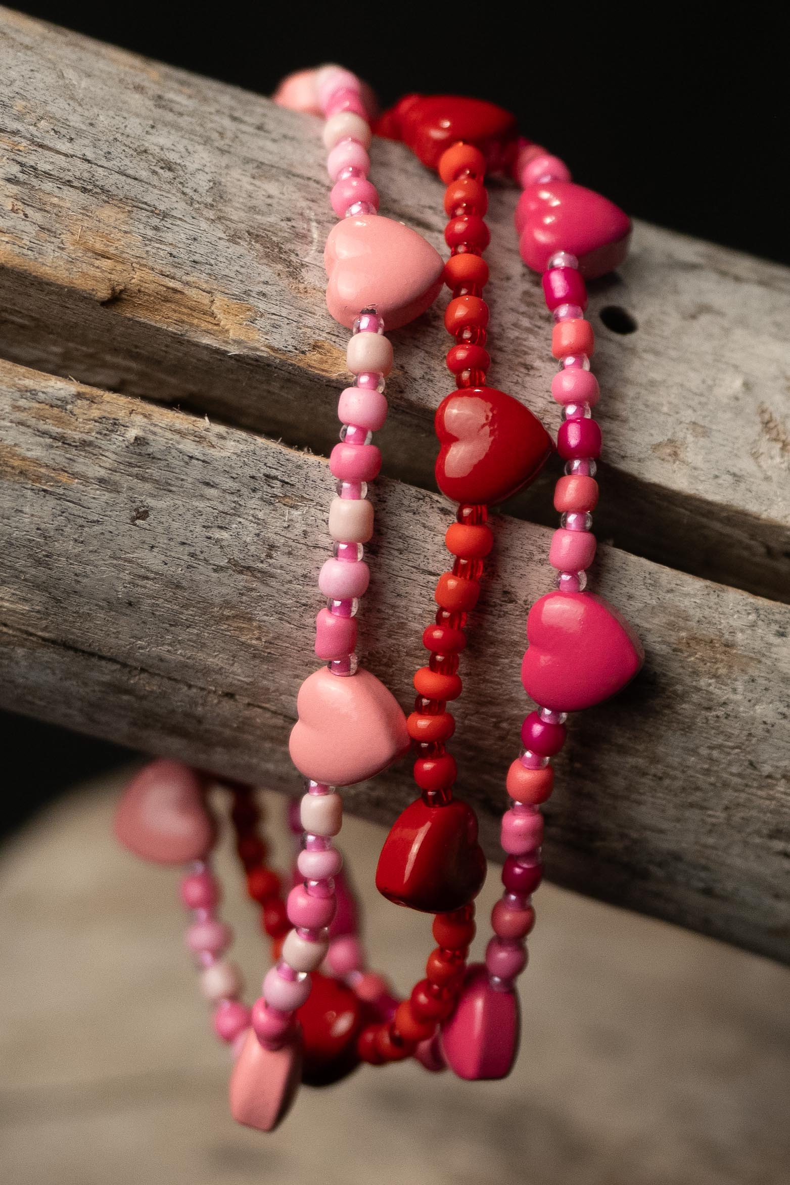 Three beaded bracelets with heart-shaped beads in red, light pink, and pink on a wooden surface.