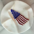 Handmade beaded American flag wind sock zipper pull with a woven blue star field and flowing red, white, and gold-tipped fringe strands, displayed on a white marble surface — Republic Zipper Pull by Amelia Heir Art Designs