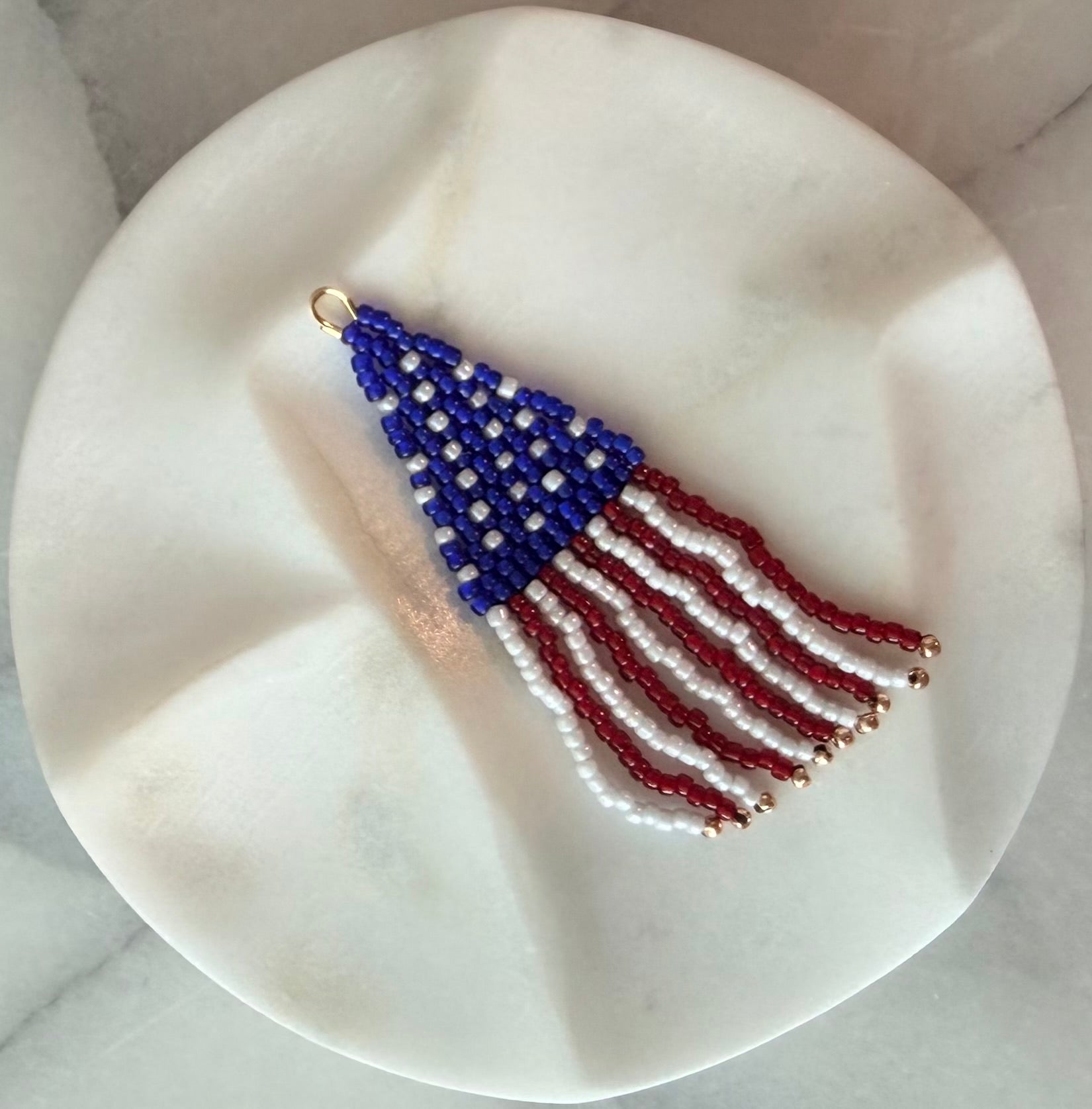 Handmade beaded American flag wind sock zipper pull with a woven blue star field and flowing red, white, and gold-tipped fringe strands, displayed on a white marble surface — Republic Zipper Pull by Amelia Heir Art Designs