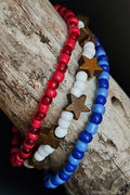 Three beaded bracelets in red, white, and blue with star-shaped wooden charms on a wooden surface.