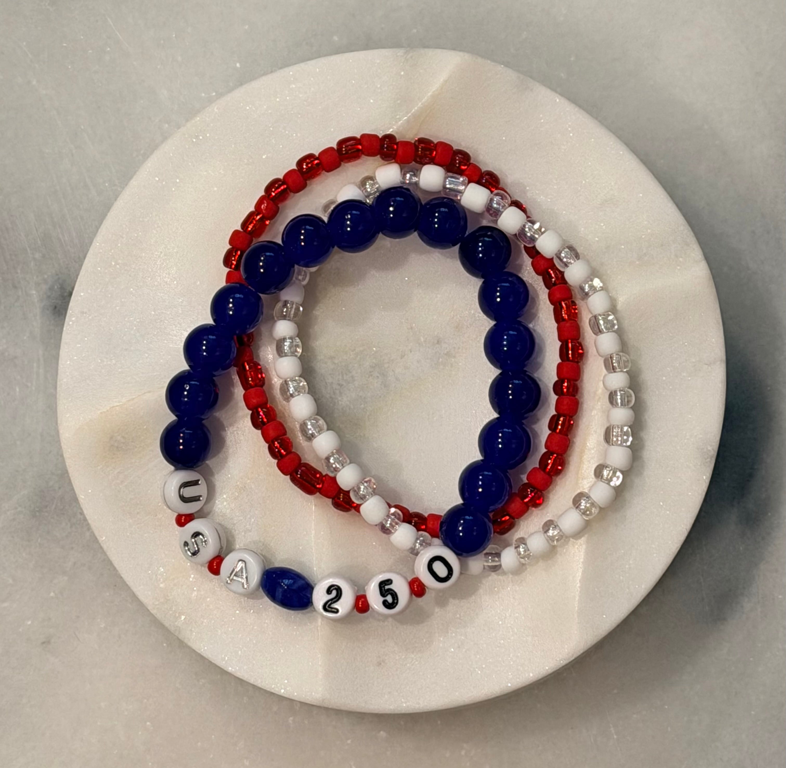 Americana Collection, 250 Anniversary, Steadfastness 250 - Three beaded bracelets in red, blue, and white with the letters USA and numbers 250 on a white marble surface, Amelia Heir Art