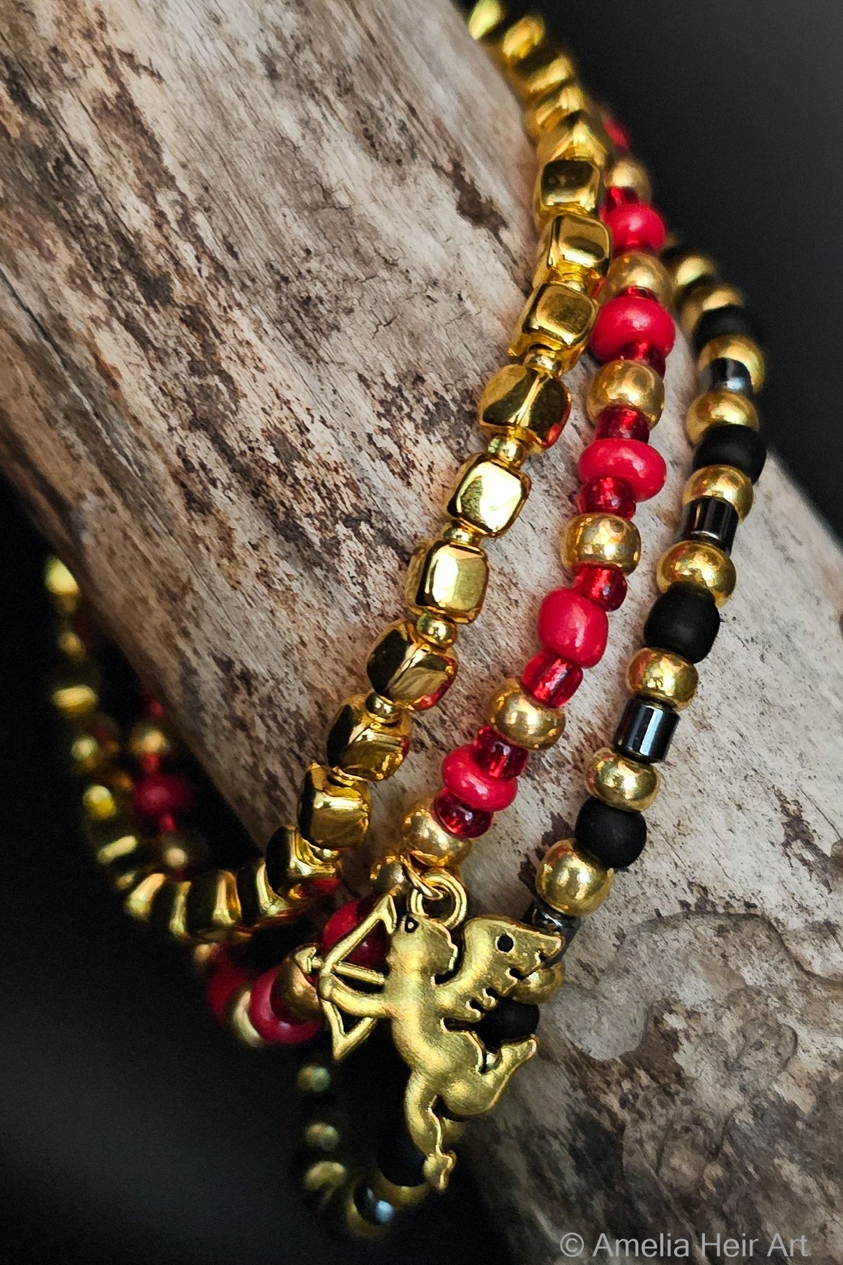 Three beaded bracelets with gold, red, and black beads and gold cupid charm on a wooden surface.