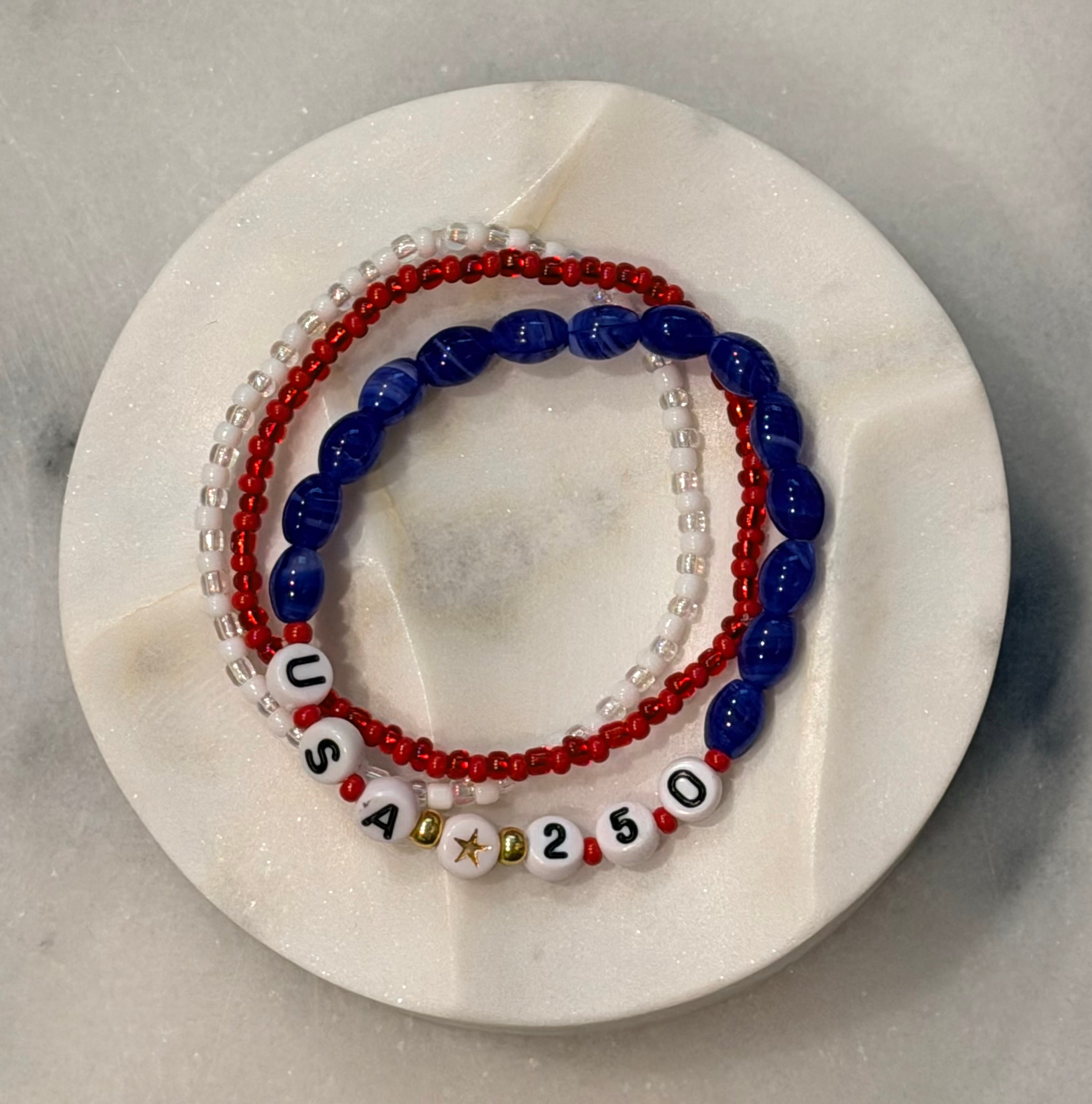 Americana Collection, 250 Anniversary, three red, white, and blue beaded bracelets with 'USA' and 250 numbers on a marble circle, Amelia Heir Art