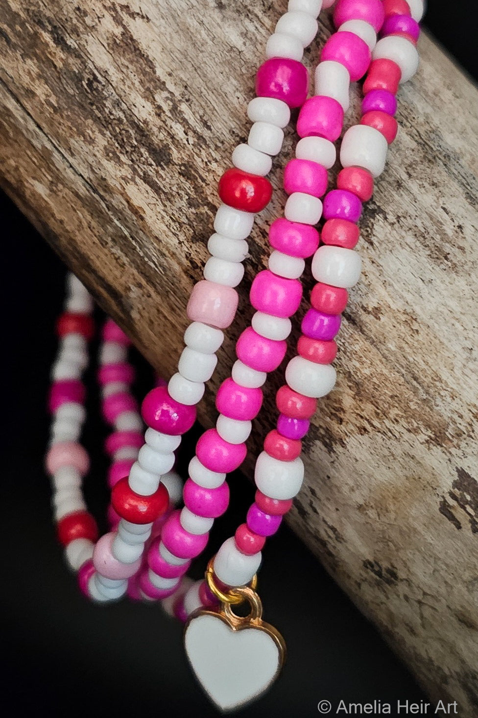 Stack of pink, white, and red beaded bracelets with a heart charm on a wooden surface.